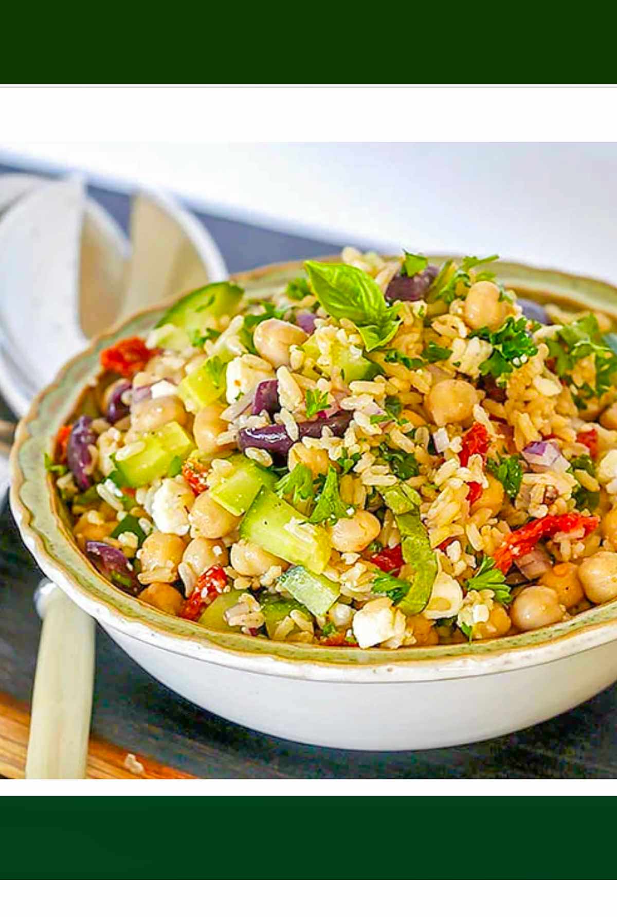 Close-up of a Mediterranean-inspired brown rice salad served in a bowl, featuring ingredients like chickpeas, cucumbers, kalamata olives, sun-dried tomatoes, feta cheese, fresh herbs, and a variety of vegetables, garnished with basil and parsley.