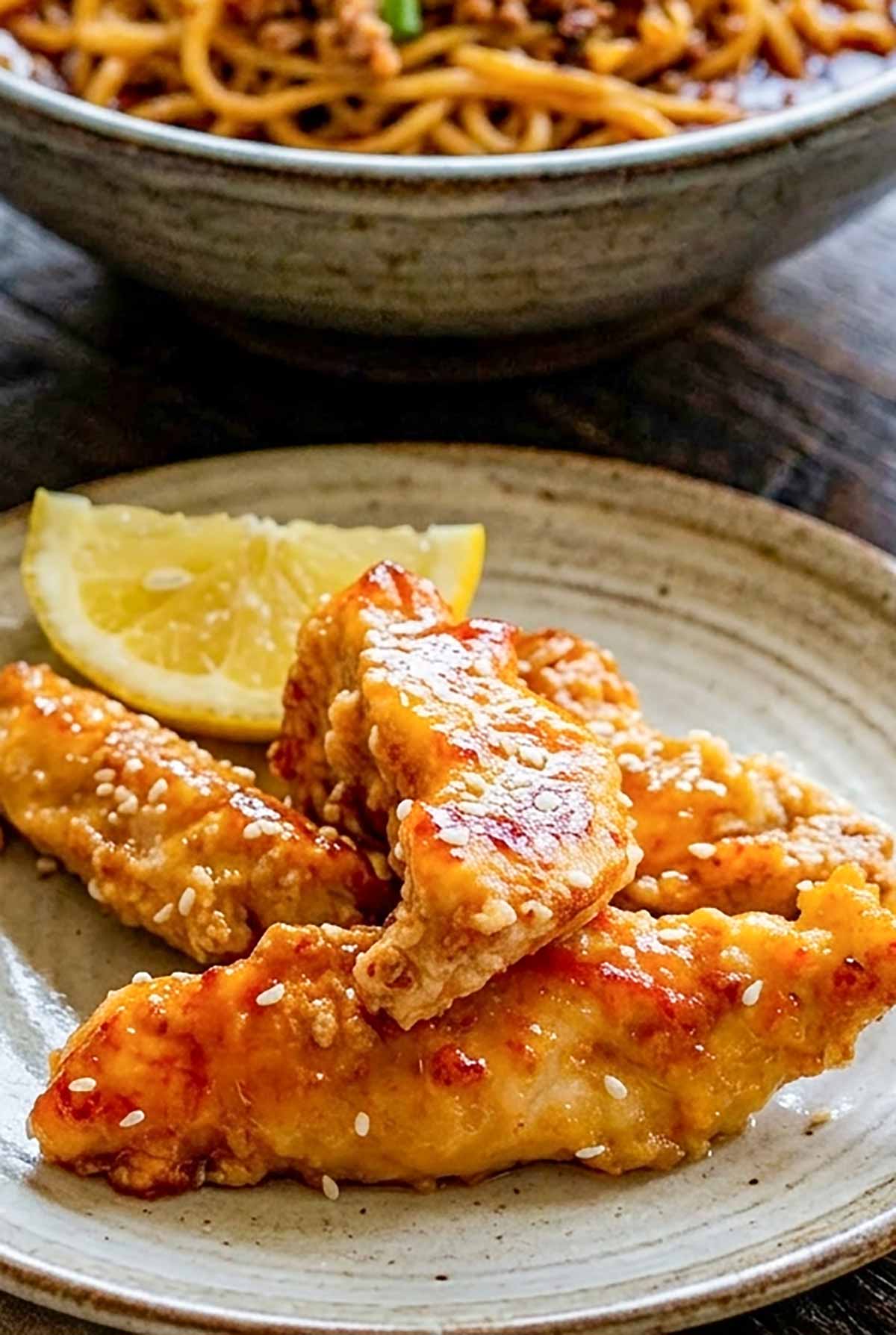 Crispy gluten free lemon chicken strips sprinkled with sesame seeds on a plate with lemon wedge