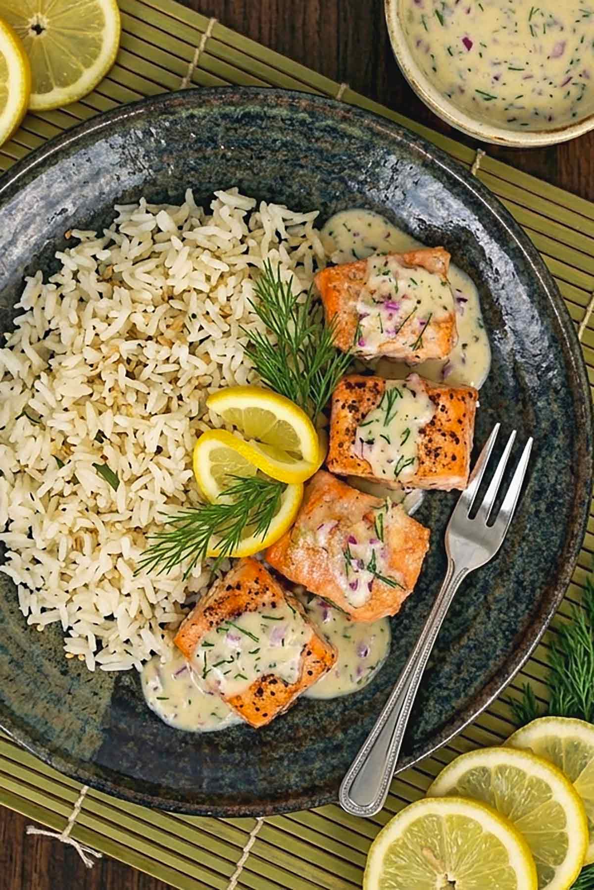 baked salmon cubes drizzles with piquant dill sauce served with a side of rice on a plate with sliced lemons