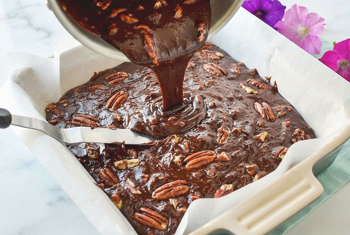 Melted chocolate with coconut milk, chopped pecans, vanilla being poured into a square pan lined with parchment paper