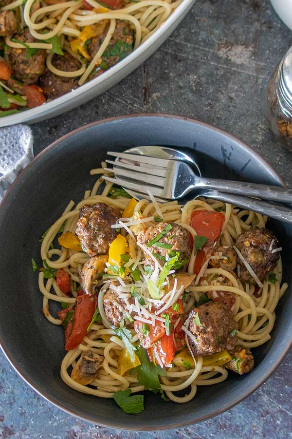 a bowl of gluten free pasta with meatballs and roasted vegetables with a fork and a spoon