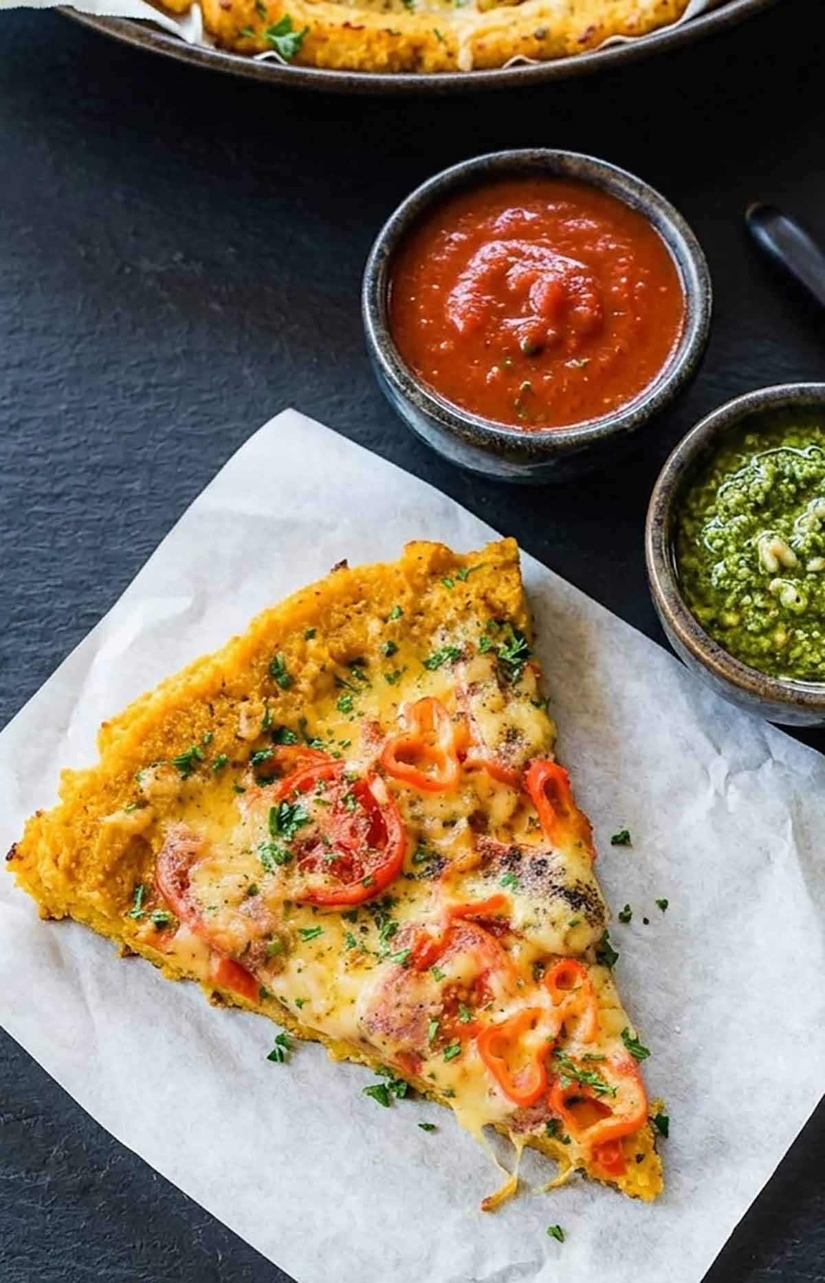 slice of butternut squash pizza crust with toppings and two dipping bowls of marinara and pesto sauce