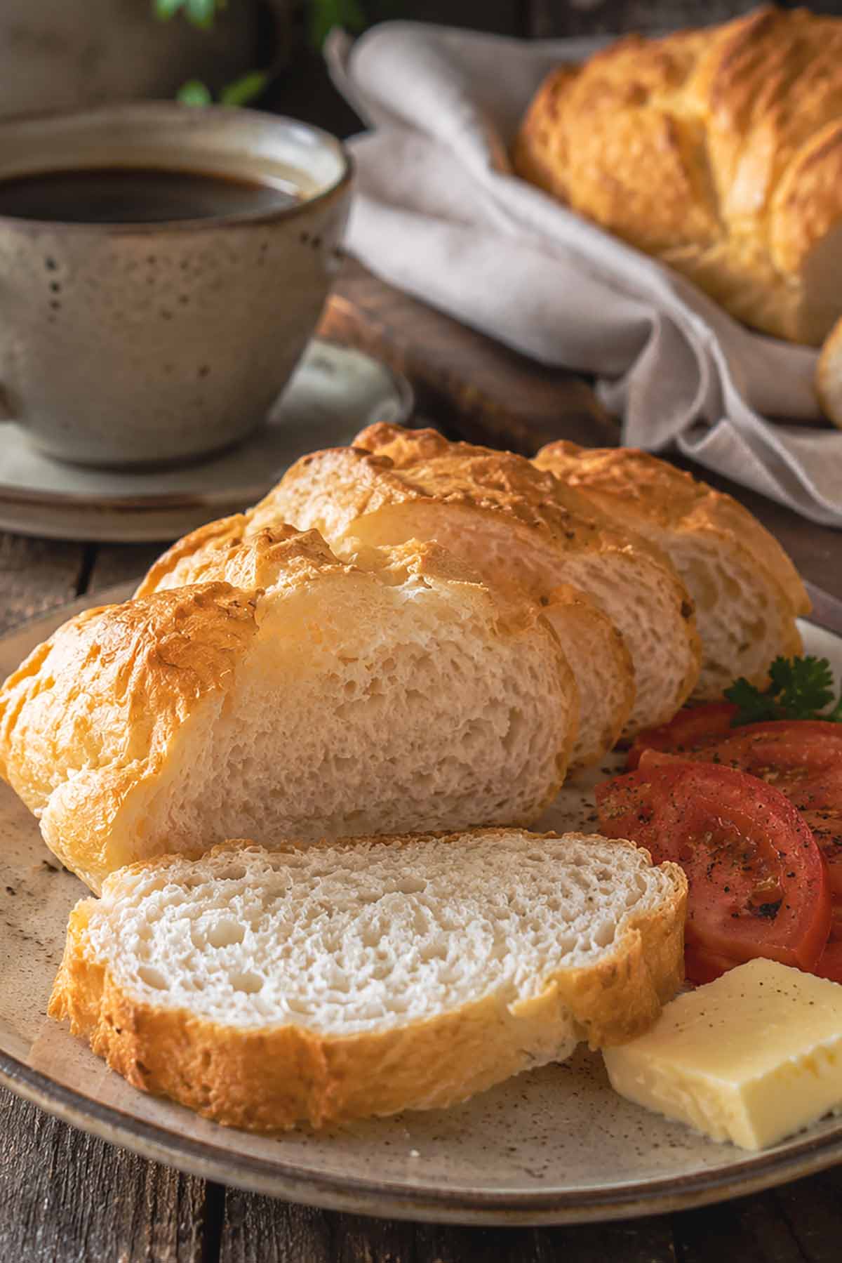 serving sliced gluten-free French bread on a plate with butter and sliced tomatoes, cup of coffee in the background