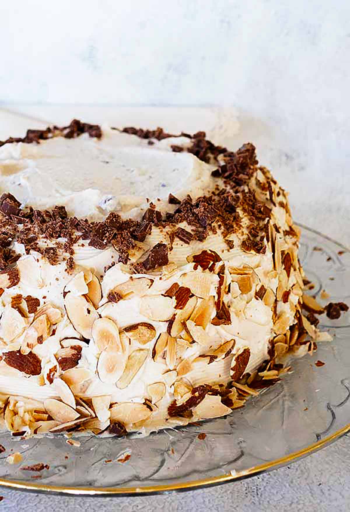 A beautiful, three-layer gluten-free cannoli cake on a cake platter, frosted with silky mascarpone cream, topped with grated chocolate and sliced almonds.