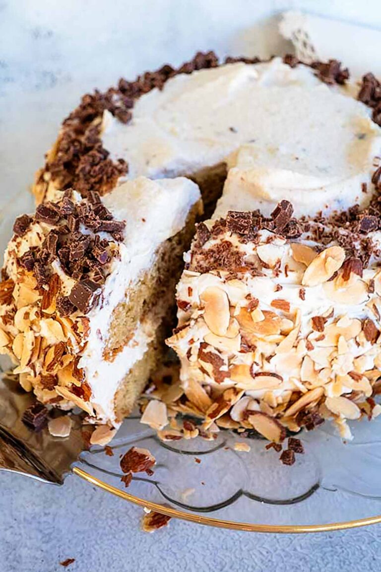 A beautiful, three-layer gluten-free cannoli cake on a glass cake platter frosted with silky mascarpone cream, and grated chocolate with sliced almonds, with a sliced piece showing the rich ricotta filling.