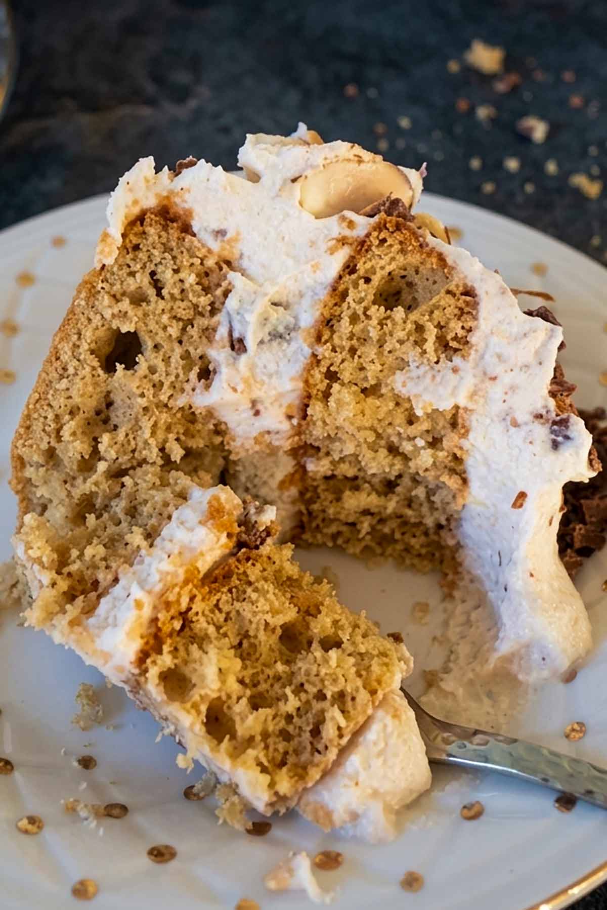 A close-up photograph of a detailed slice of multi-layer sponge cake with white frosting, almond slivers, and finely chopped chocolate, served on a white patterned, gold-rimmed plate