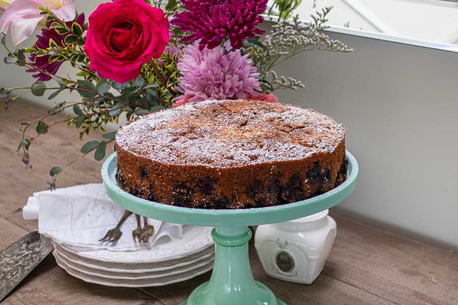 whole snacking blueberry cake on a cake platter dusted with powdered sugar with desert plates and forks