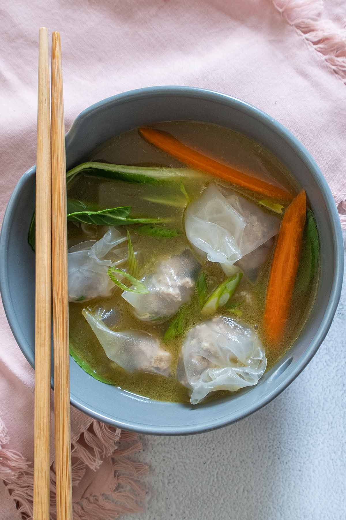wonton soup with dumplings, bok choy and carrots in a bowl with chopsticks