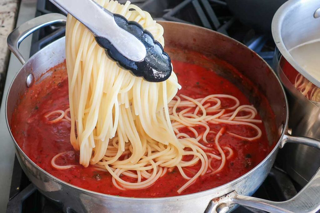 adding cooked pasta with tongs into the sauce in a skillet