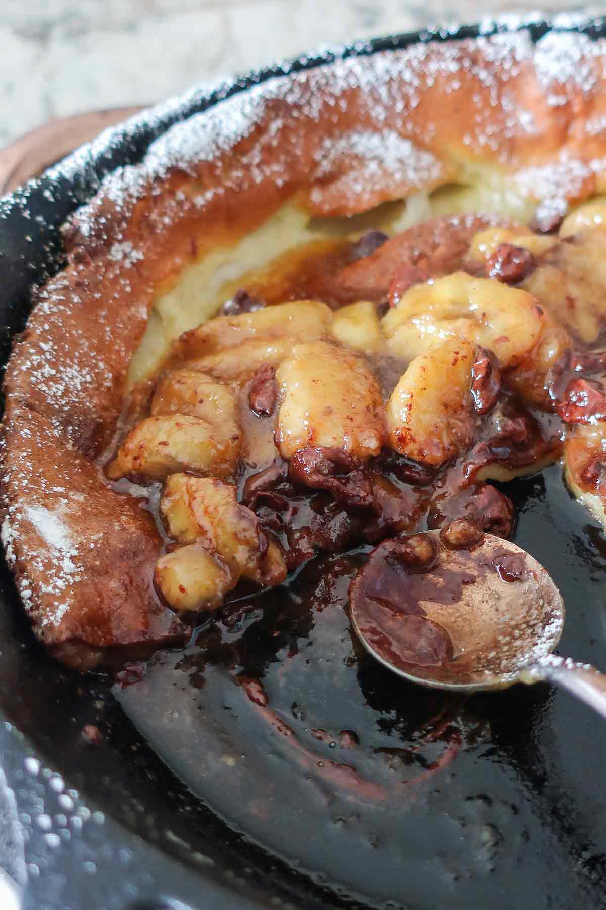 a spoon in a skillet scooping out dutch pancake with banan chocolate sauce