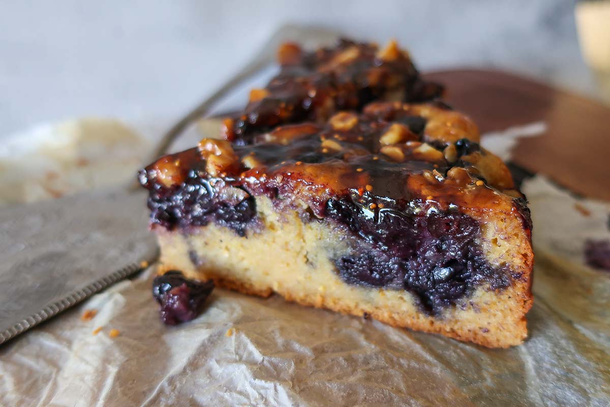 Close-up of a slice of cornmeal cake on a dessert plate, studded with juicy blueberries and topped with a glossy, buttery fruit glaze