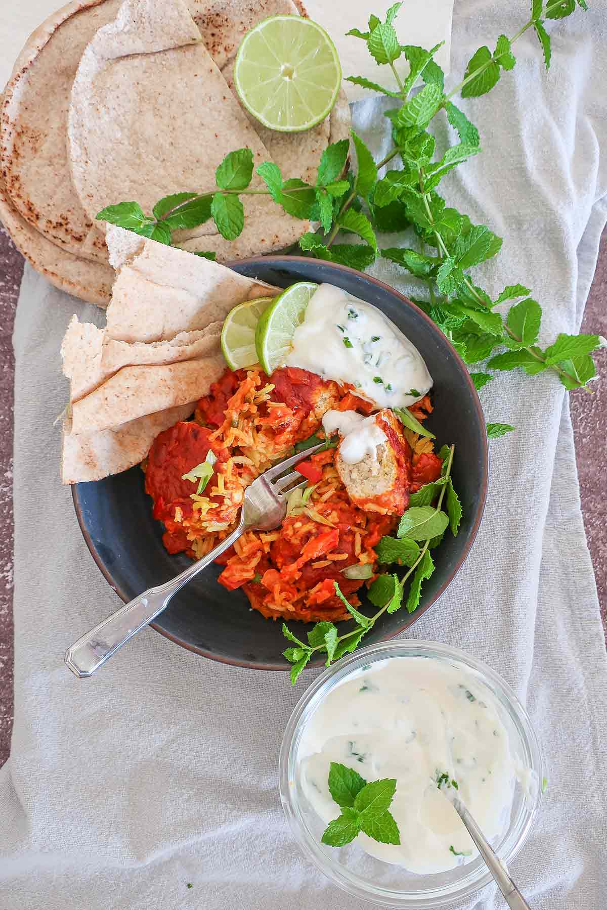 top view of a bowl with butter chicken meatballs, rice, sauce, yogurt, herbs, Indian bread