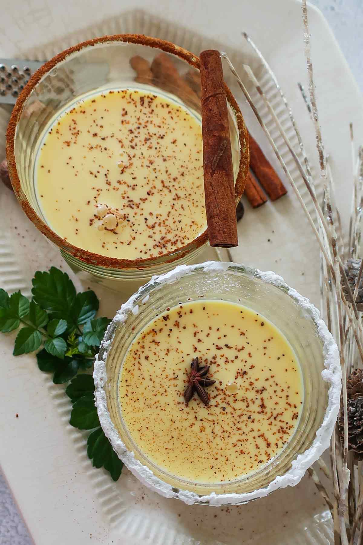 two glasses of eggnog, rimmed with cinnamon and sugar, one garnished with a cinnamon stick, the other features a star anise