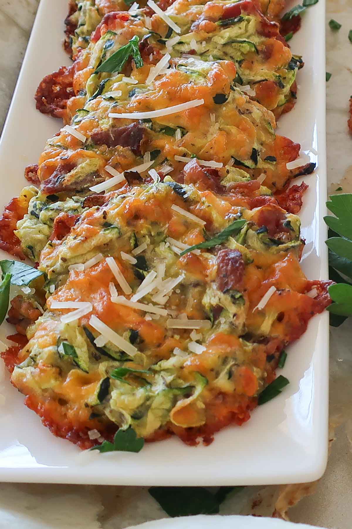 A row of cheddar bacon zucchini bites on a plate, topped with fresh parsley and grated Parmesan cheese.
