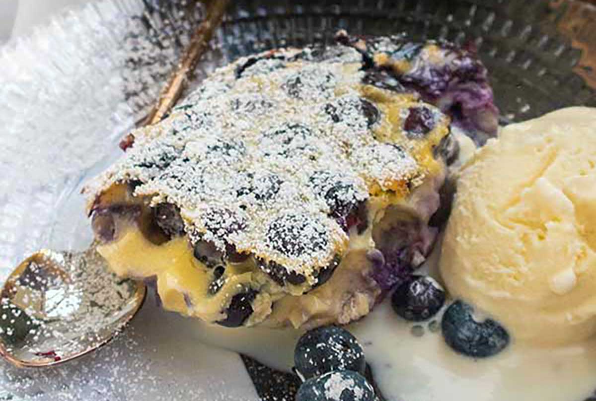 A slice of easy blueberry clafoutis with a custard-like texture, served warm on a plate alongside a scoop of creamy vanilla ice cream—simple, elegant