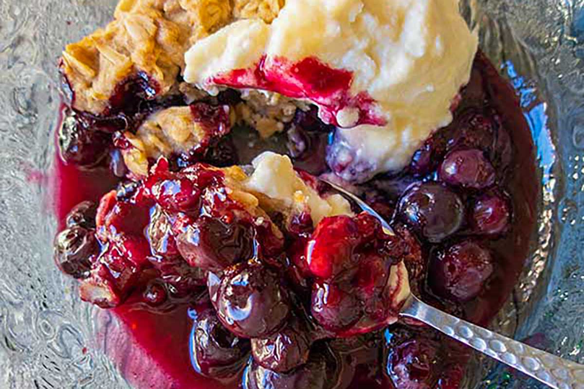Warm blueberry crumble served in a glass bowl, overflowing with juicy blueberry sauce and topped with a creamy scoop of vanilla ice cream slowly melting into the fruit—comfort dessert at its finest.