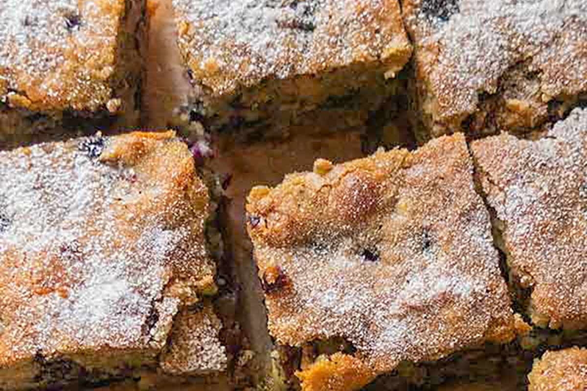 Blueberry breakfast cake sliced into squares and lightly dusted with powdered sugar, displayed on a rustic wooden board—moist, golden, and studded with juicy blueberries