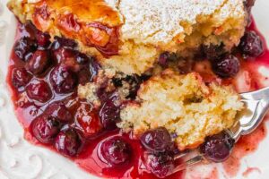 Blueberry pudding cake served on a white plate with a fork revealing its juicy, fruit-filled center. The moist texture and vibrant purple-blue berries are visible in on the fork
