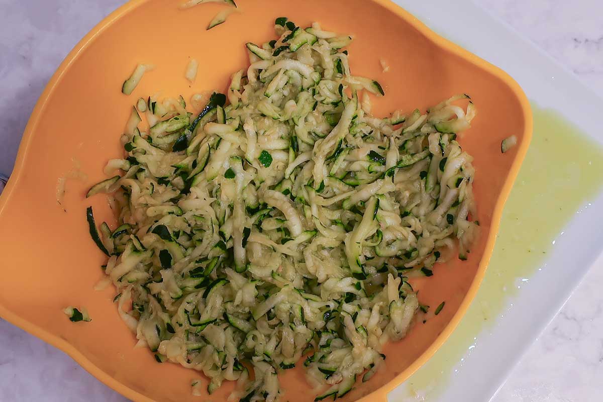 Shredded zucchini draining in a colander, with a plate underneath catching the excess liquid.