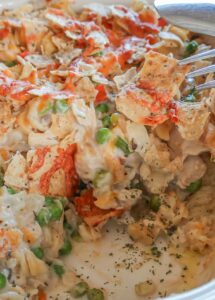 Baked gluten-free chicken noodle casserole in a deep ceramic casserole dish, showing creamy sauce, chunks of chicken, carrots, peas, and noodles; one slice is missing, revealing the soft, cheesy interior.