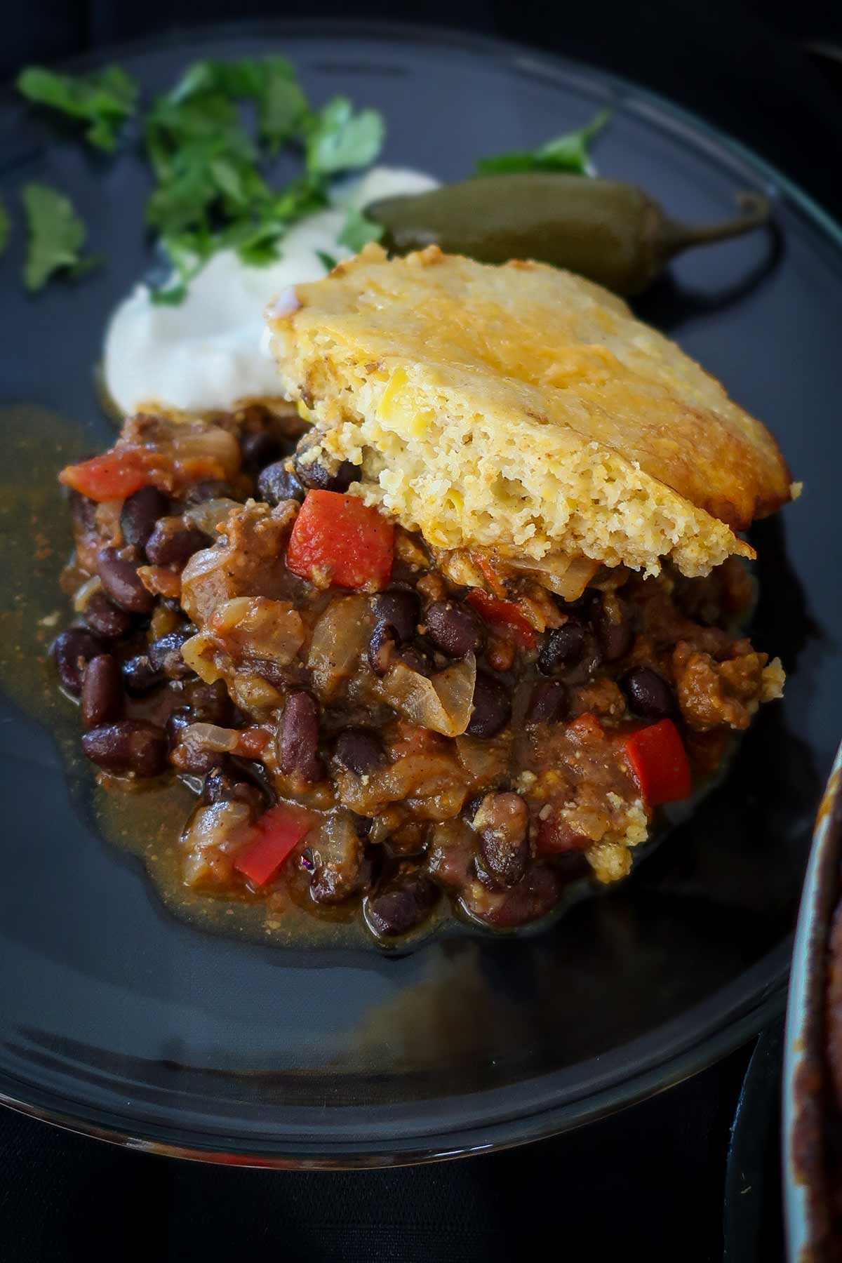 serving of pumpkin chilli topped with buttermilk cornbread on a plate with sour cream, cilantro and jalapeño