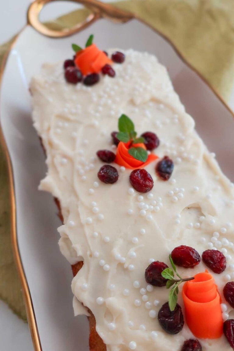 Gluten-free carrot honey pound cake topped with cream cheese frosting, dried cranberries, carrot ribbon decorations, white sugar pearls, and fresh mint leaves on a serving tray.