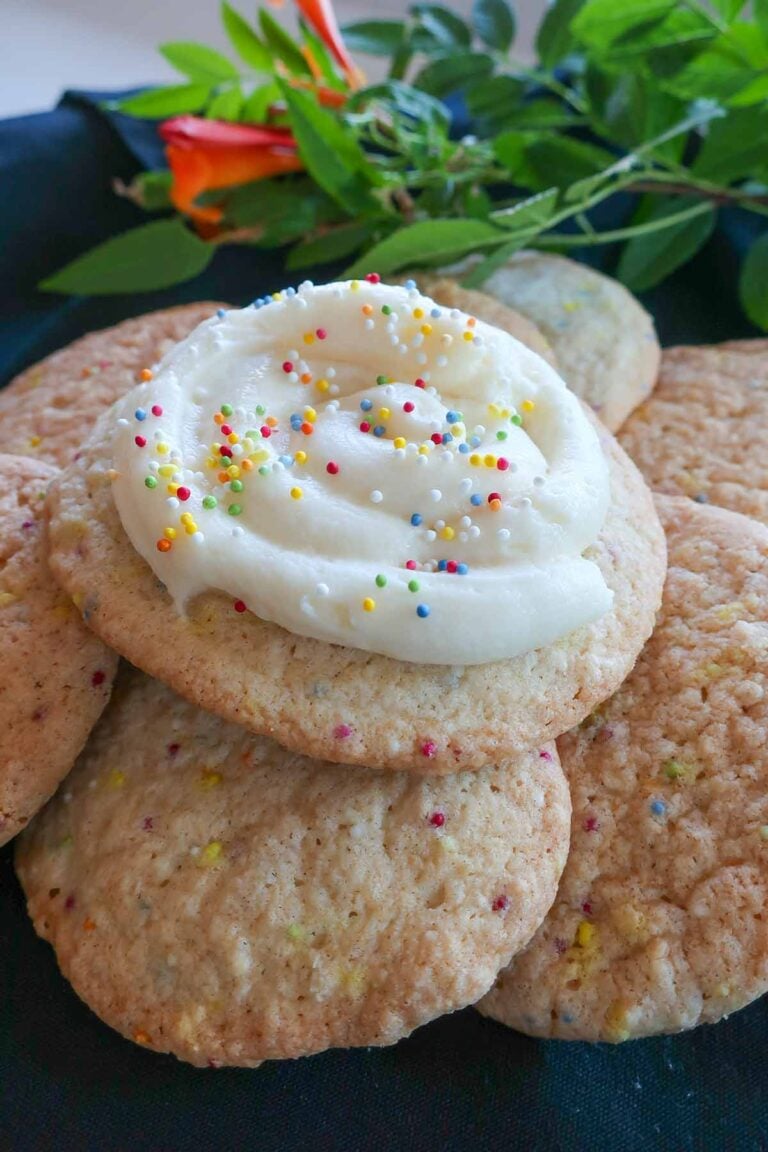 Stack of gluten-free crumbs-style sugar cookies topped with swirled cream cheese frosting and colorful sprinkles, with greenery in the background.