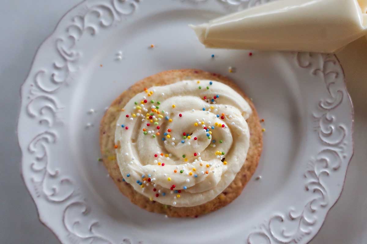Soft Crumbl sugar cookie topped with swirled cream cheese frosting and colorful sprinkles on a white plate