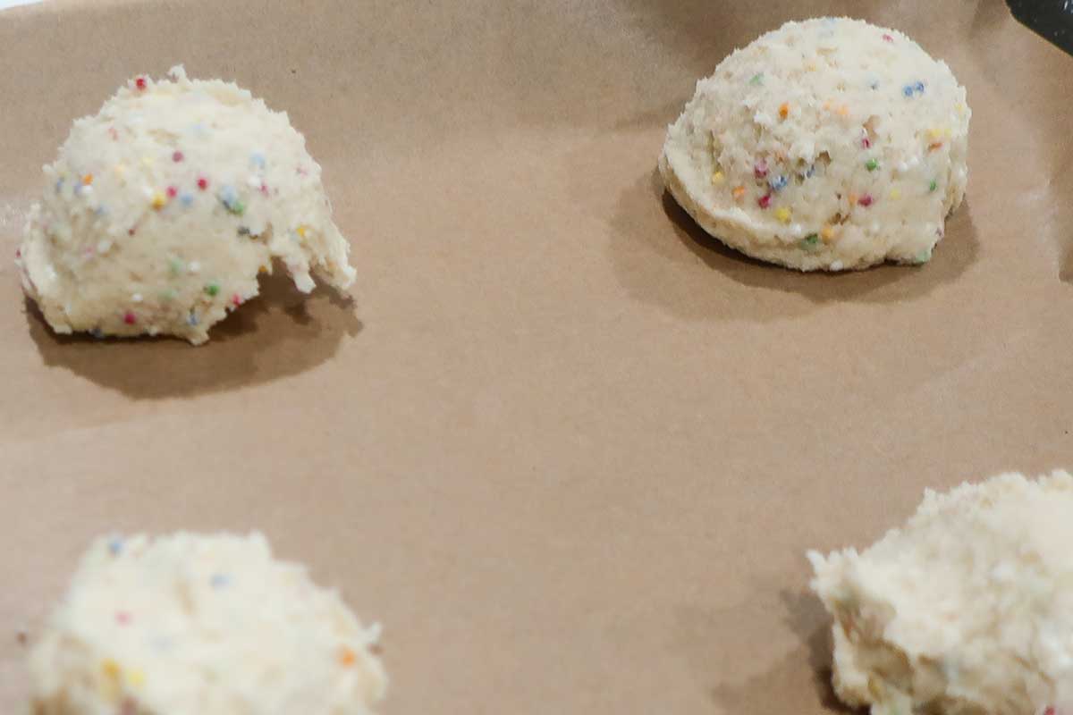 Scoops of unbaked funfetti sugar cookie dough with colorful sprinkles on a parchment-lined baking sheet.