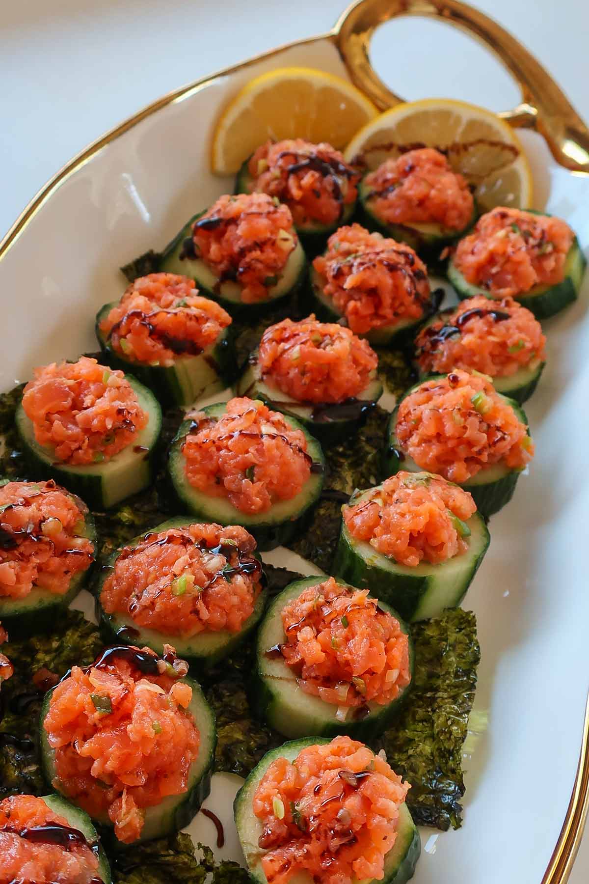 close up of Cucumber and smoked salmon bites arranged on white 3x12 inch platter trimmed in gold with thin lemon slices