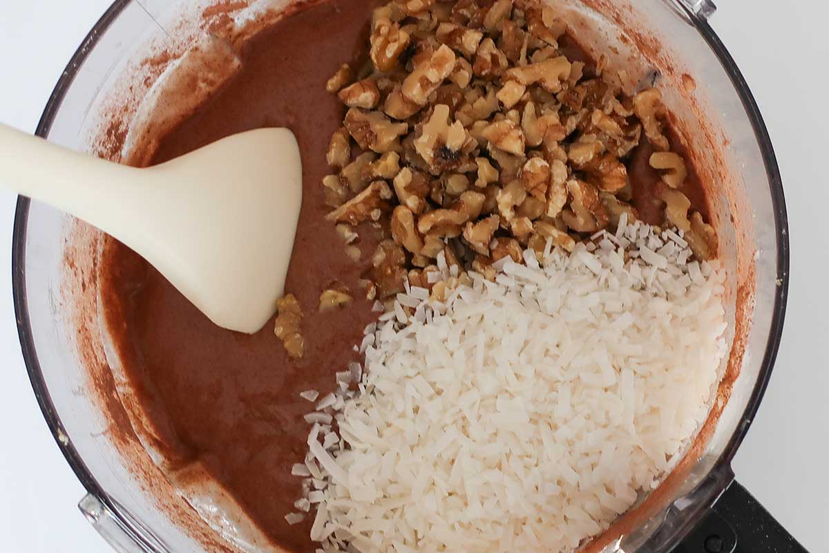 Folding shredded coconut and chopped walnuts into chocolate cake batter with a spatula in a food processor bowl