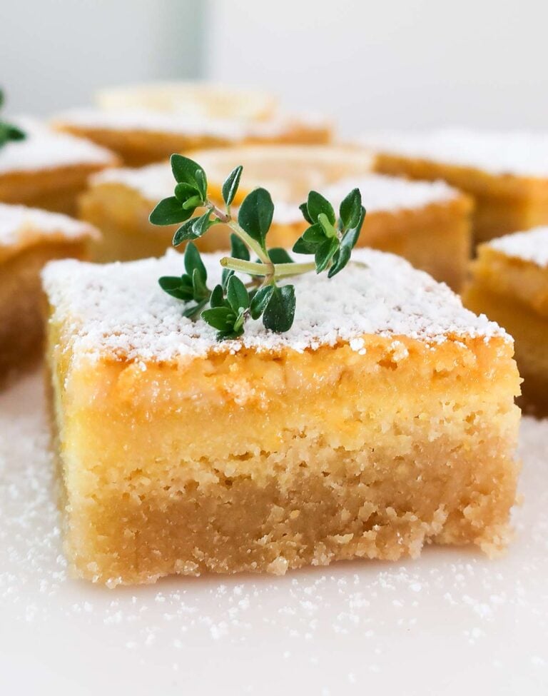 A single square lemon bar topped with white powdered sugar and a small green herb garnish, with other blurred lemon bars in the background on a white surface.