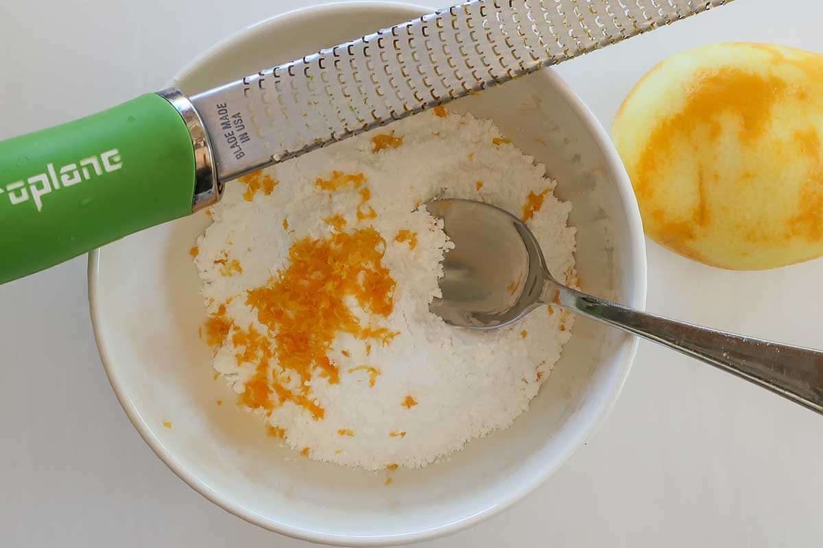 A close-up of fresh lemon zest being rubbed into white powdered sugar to release aromatic essential oils.