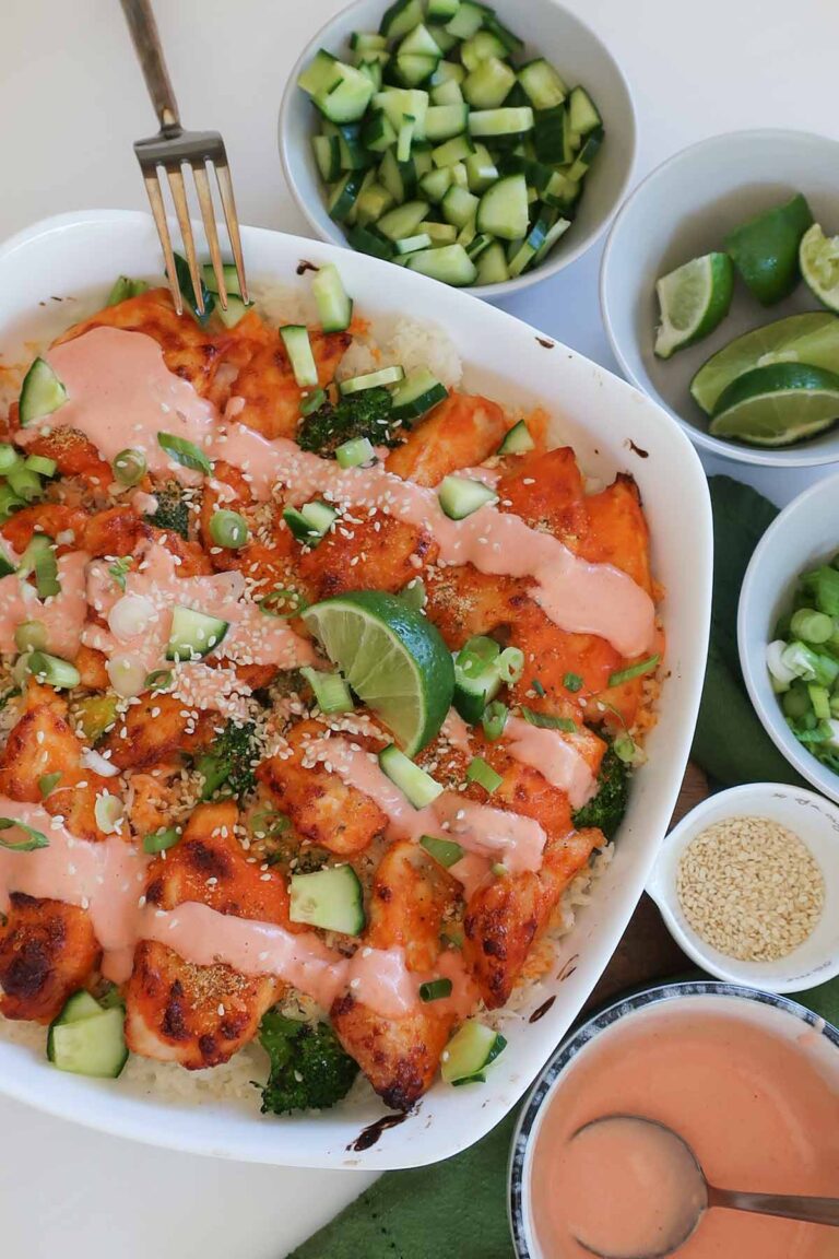 GF Bang Bang chicken and rice casserole in a baking dish topped with creamy sauce, cucumber, green onions, and sesame seeds, served with small bowls of extra fresh toppings on the side.