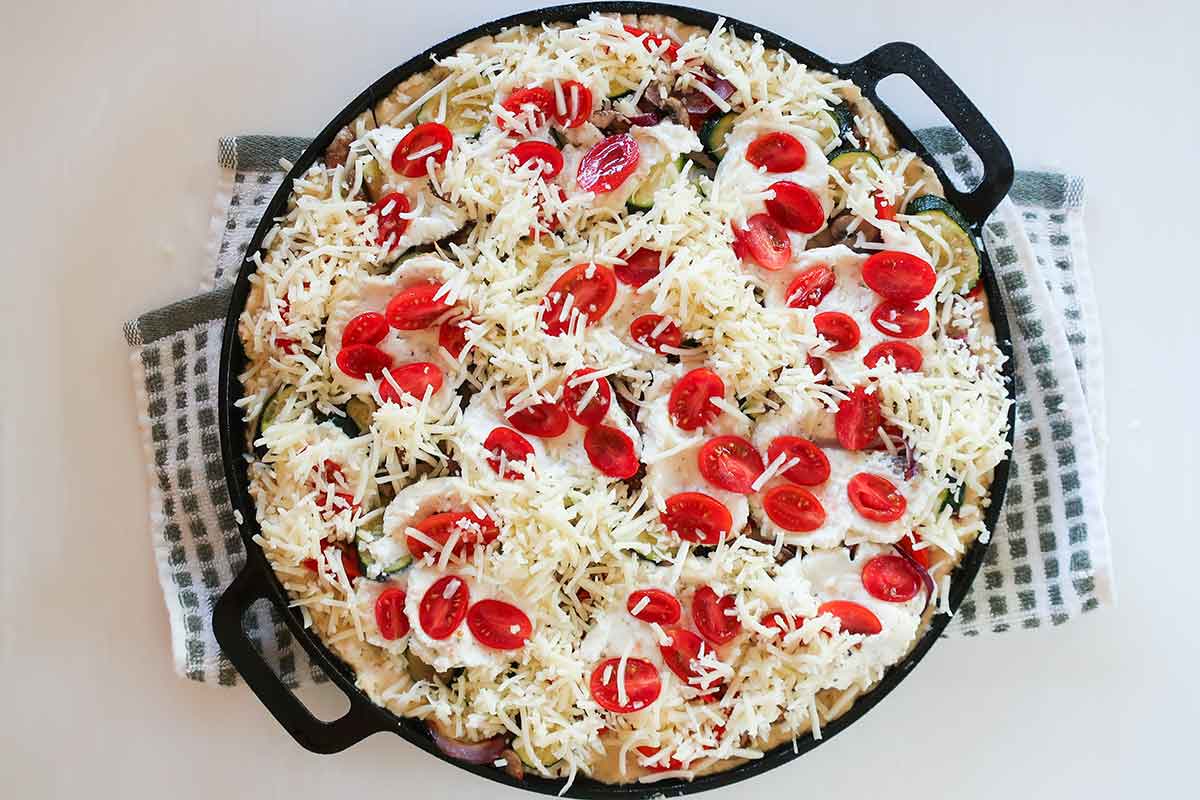 Unbaked gluten-free cast iron pizza topped with roasted vegetables, ricotta, halved cherry tomatoes, and cheese