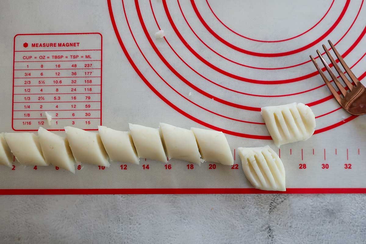 One piece of gnocchi dough rolled into a long rope, alongside small cut dough pillows with traditional fork ridge details on a countertop.