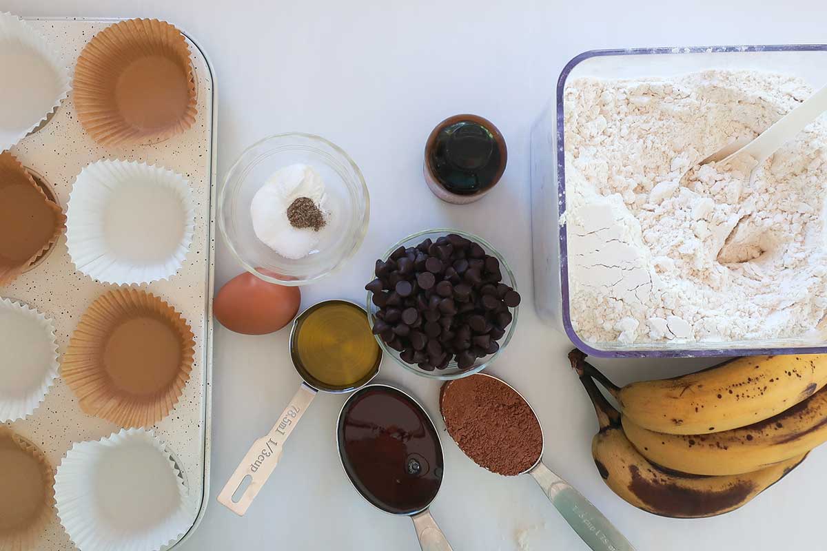 Ingredients on the counter, flour, bananas, egg, maple syrup, cardamom, cocoa powder, choc chips, olive oil