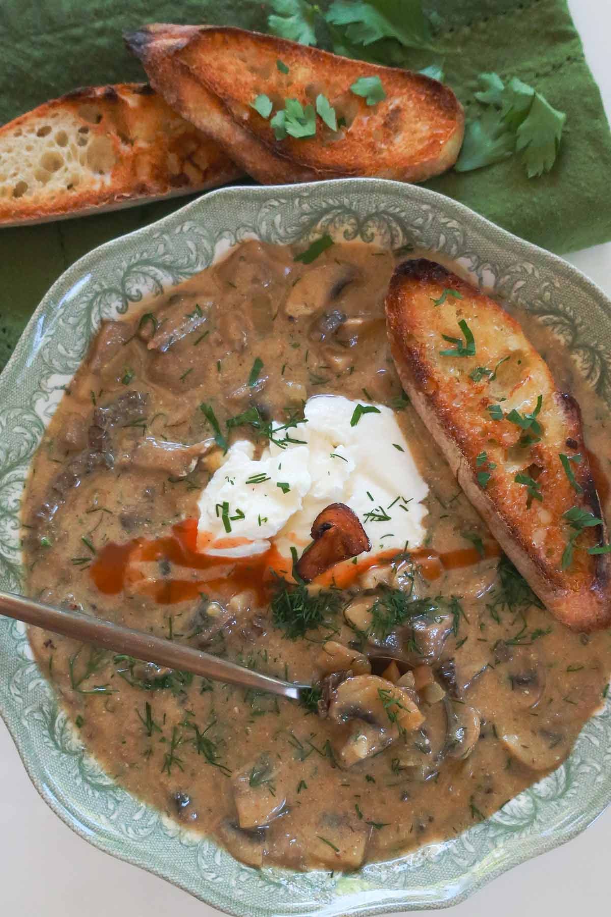 A bowl of authentic Hungarian thick and creamy mushrooms soup topped with sour cream crusty GF bread, fresh dill and spicy sauce