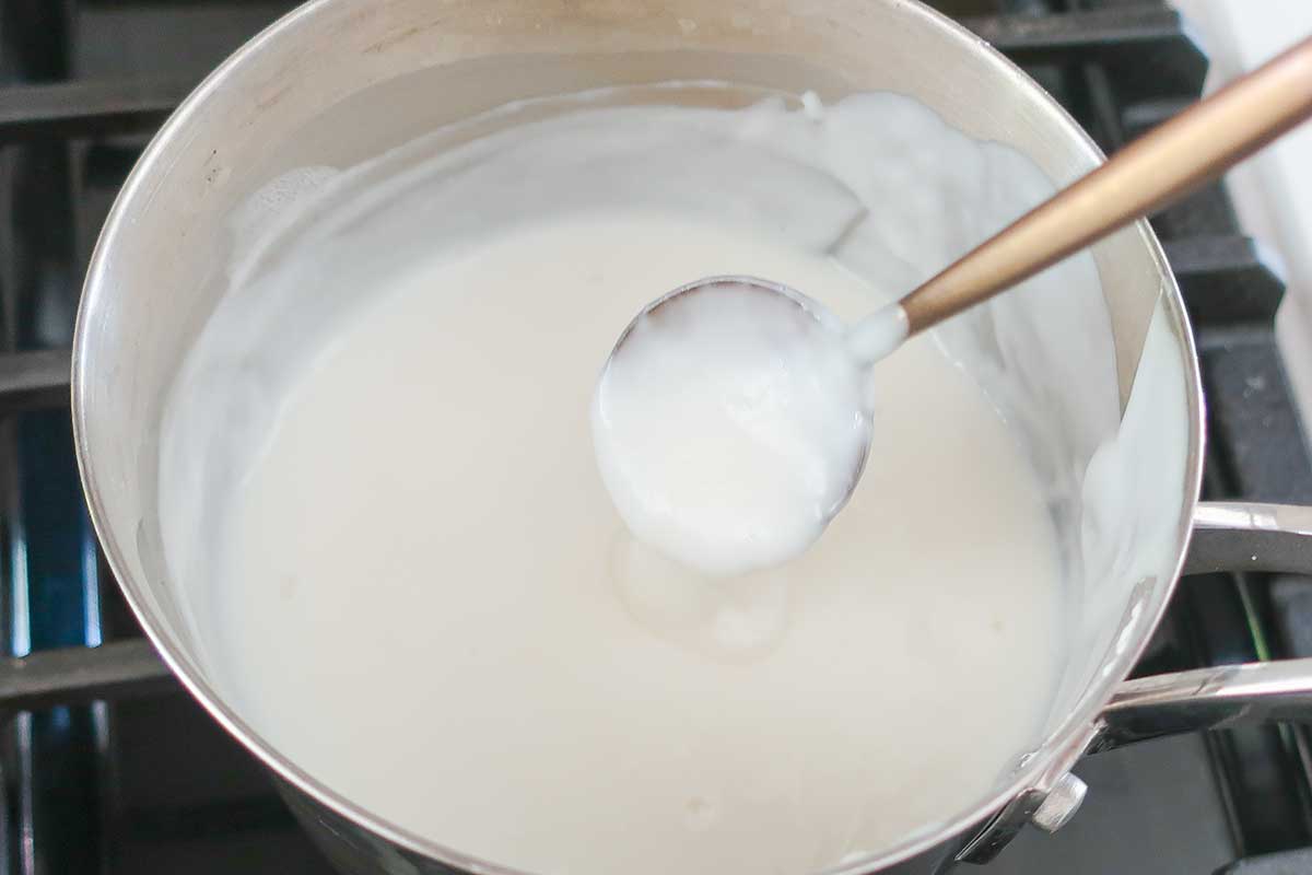 A close-up view of a metal pot on a stove burner filled with a very thick, heavy, and pale cooked milk and starch pudding base for Ermine frosting. A spoon coated in the dense mixture is submerged in the center.