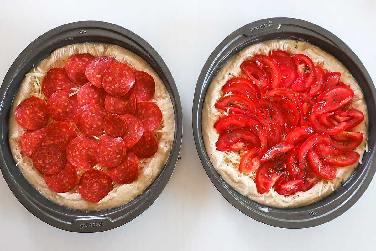 two 9" pizzas before baking one topped with pepperoni one with fresh tomatoes in round baking pans
