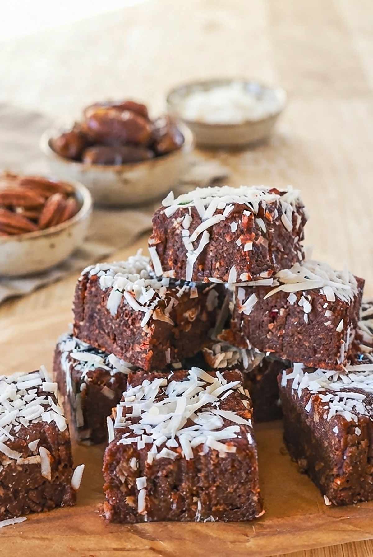 close up of Fudgy No-Bake Grain-Free Chocolate Date Bars with 3 bowls in the background of dates, pecans and shredded coconut